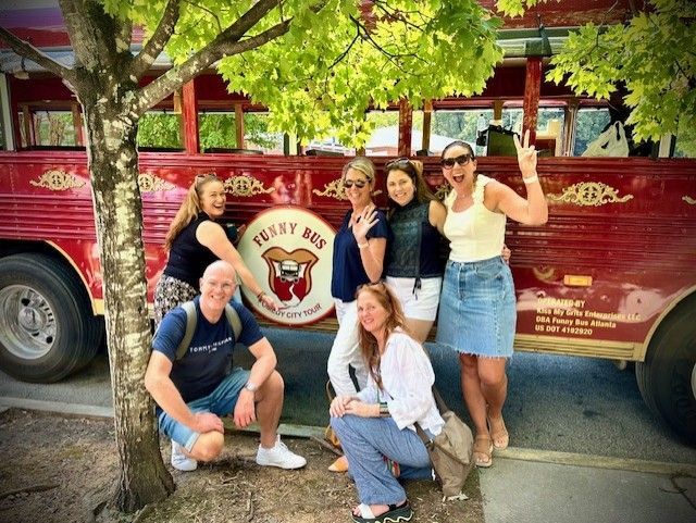 Six people pose for a photo in front of a red vintage-style tour bus parked near a tree.
