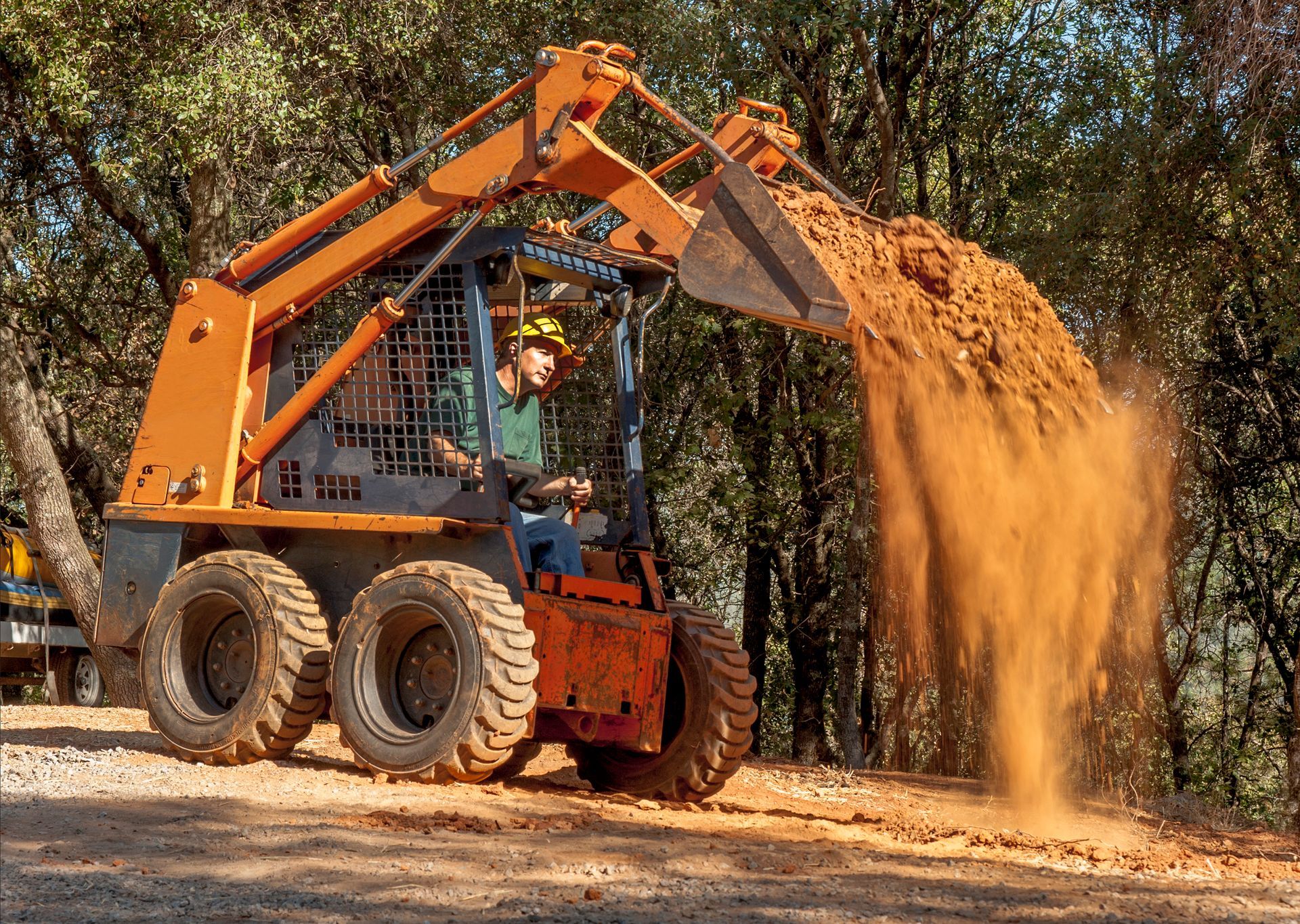 An orange skid steer dumps a load of red dirt.