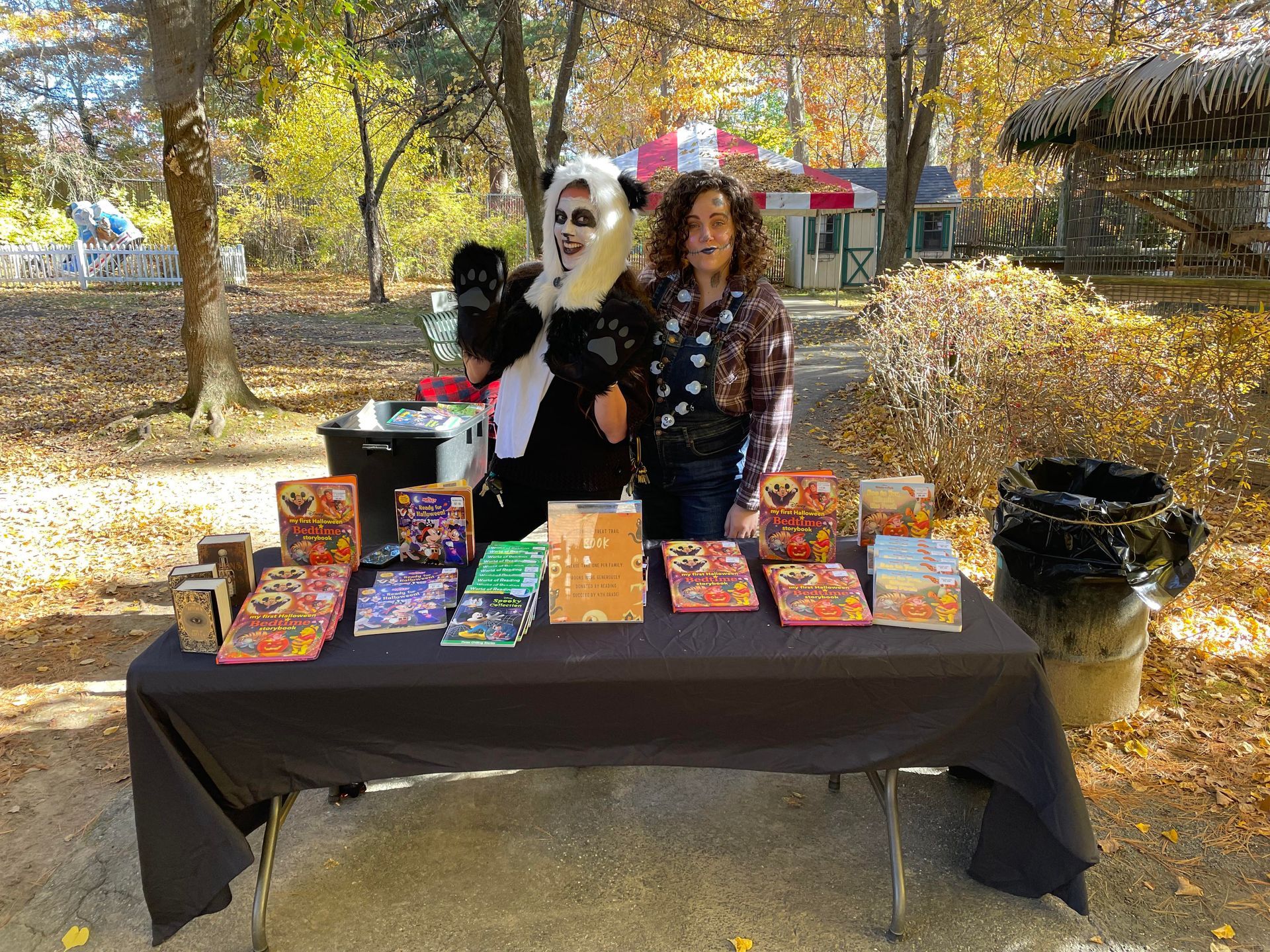 Two Zoo staff members dressed as a panda and a scarecrow pose behind a table of free books at Spooky Safari.