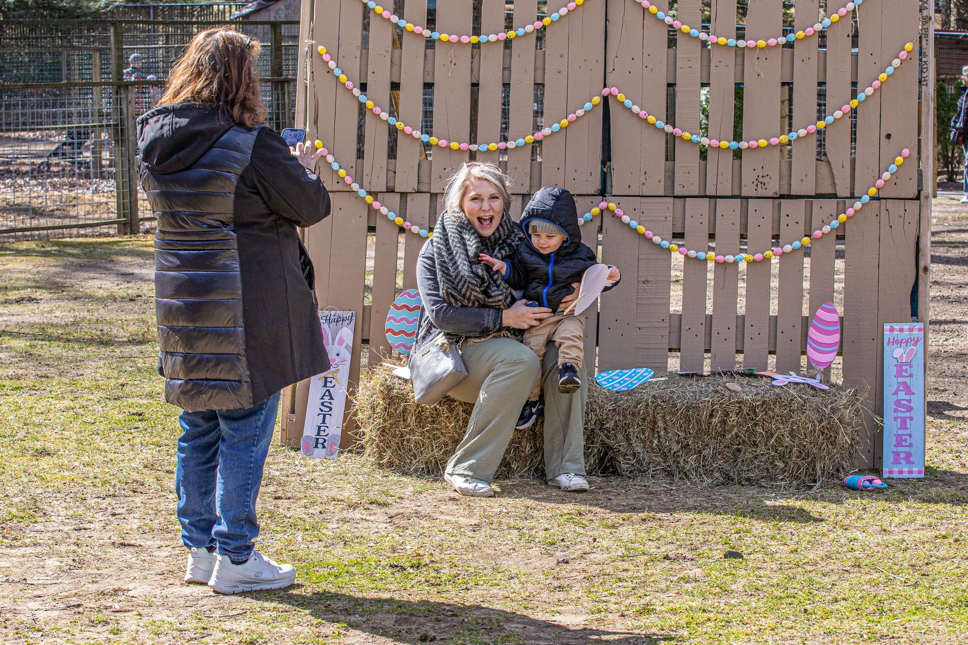 A woman in a coat takes a picture of a woman and a child sitting on a bale of hay in front of a photo backdrop.