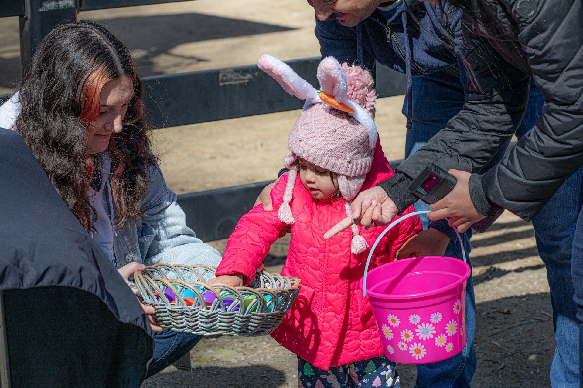 A child in a pink coat and wearing bunny ears reaches into a basket to select a plastic egg.
