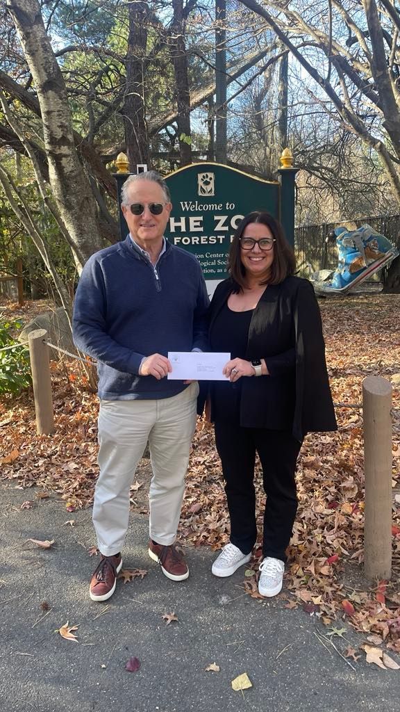 A man stands presenting a check to a woman in front of The Zoo in Forest Park's welcome sign.