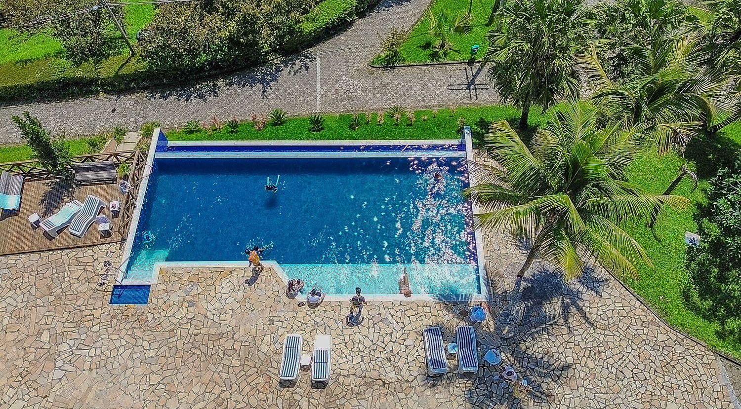Vista aérea de uma piscina retangular com pessoas nadando e relaxando, cercada por um pátio de pedra e vegetação.