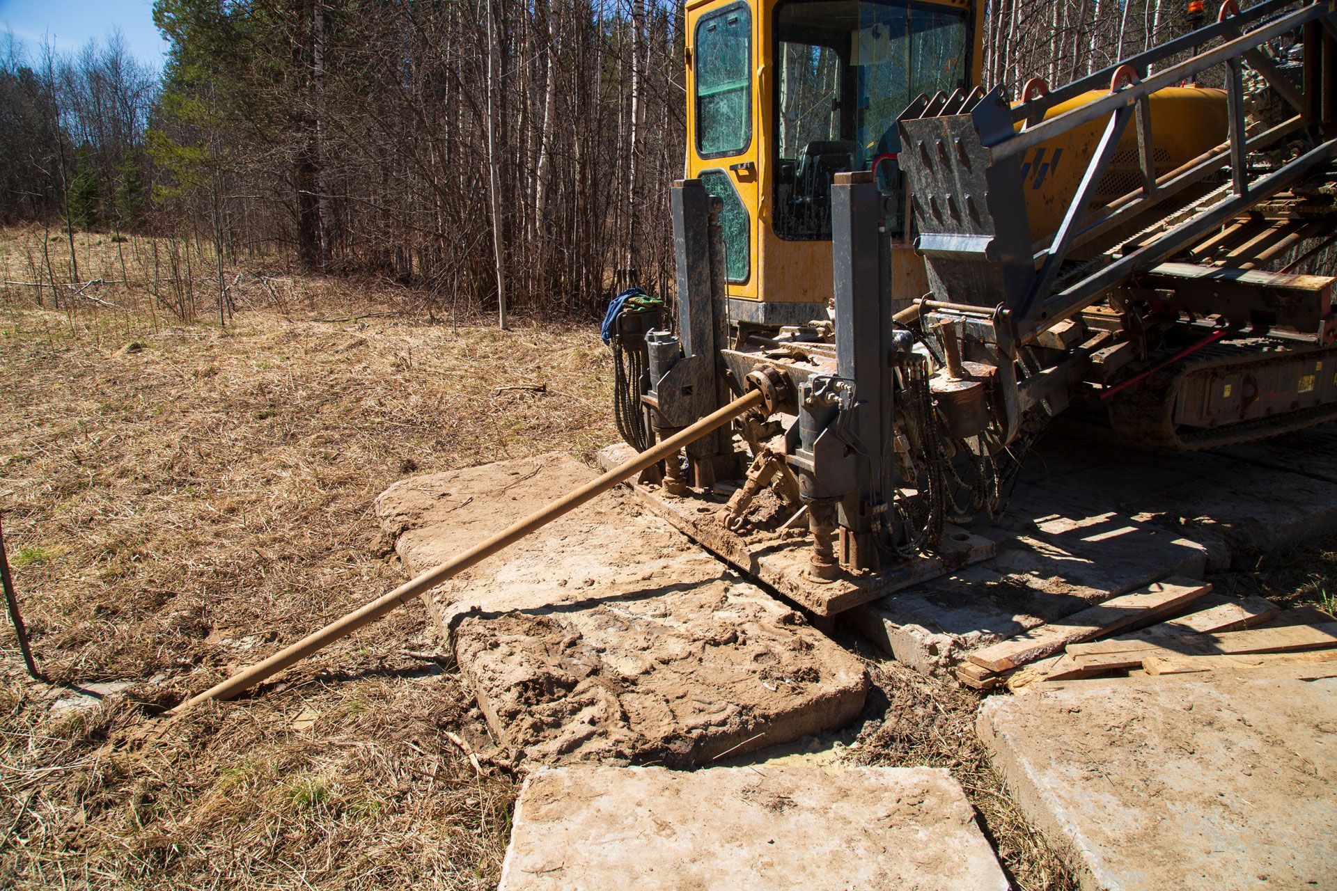 Yellow drilling machine bores into the ground surrounded by dry grass, wood boards, and trees.
