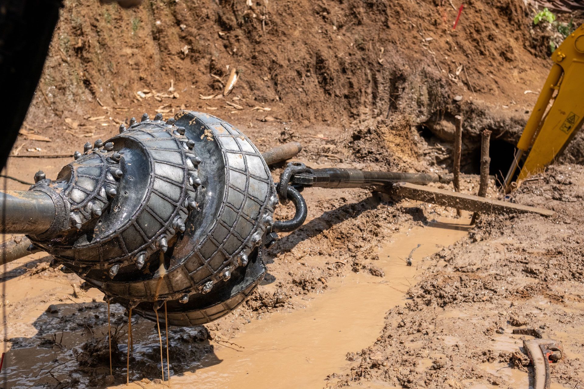 A large boring machine, with circular cutting heads, is emerging from a muddy trench.