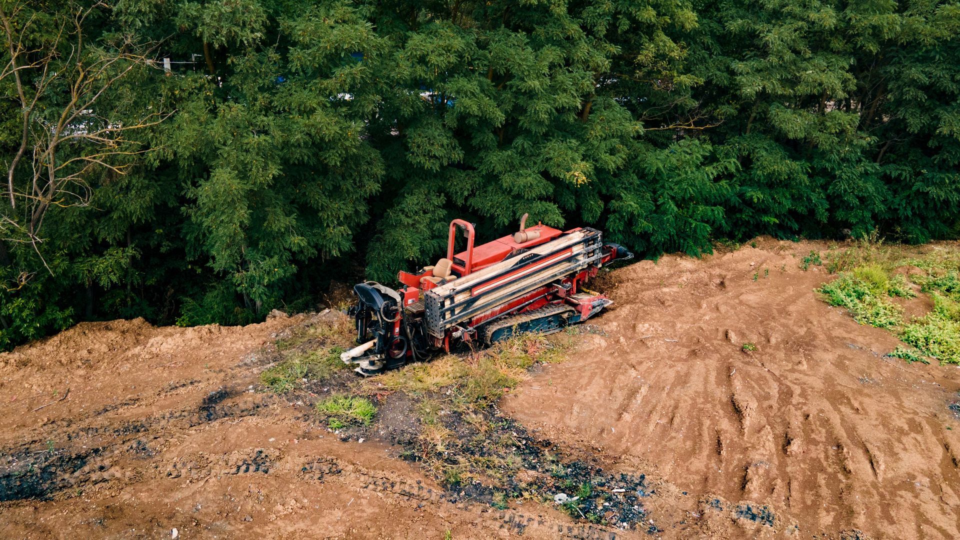 Red drilling machine on muddy ground near green trees.