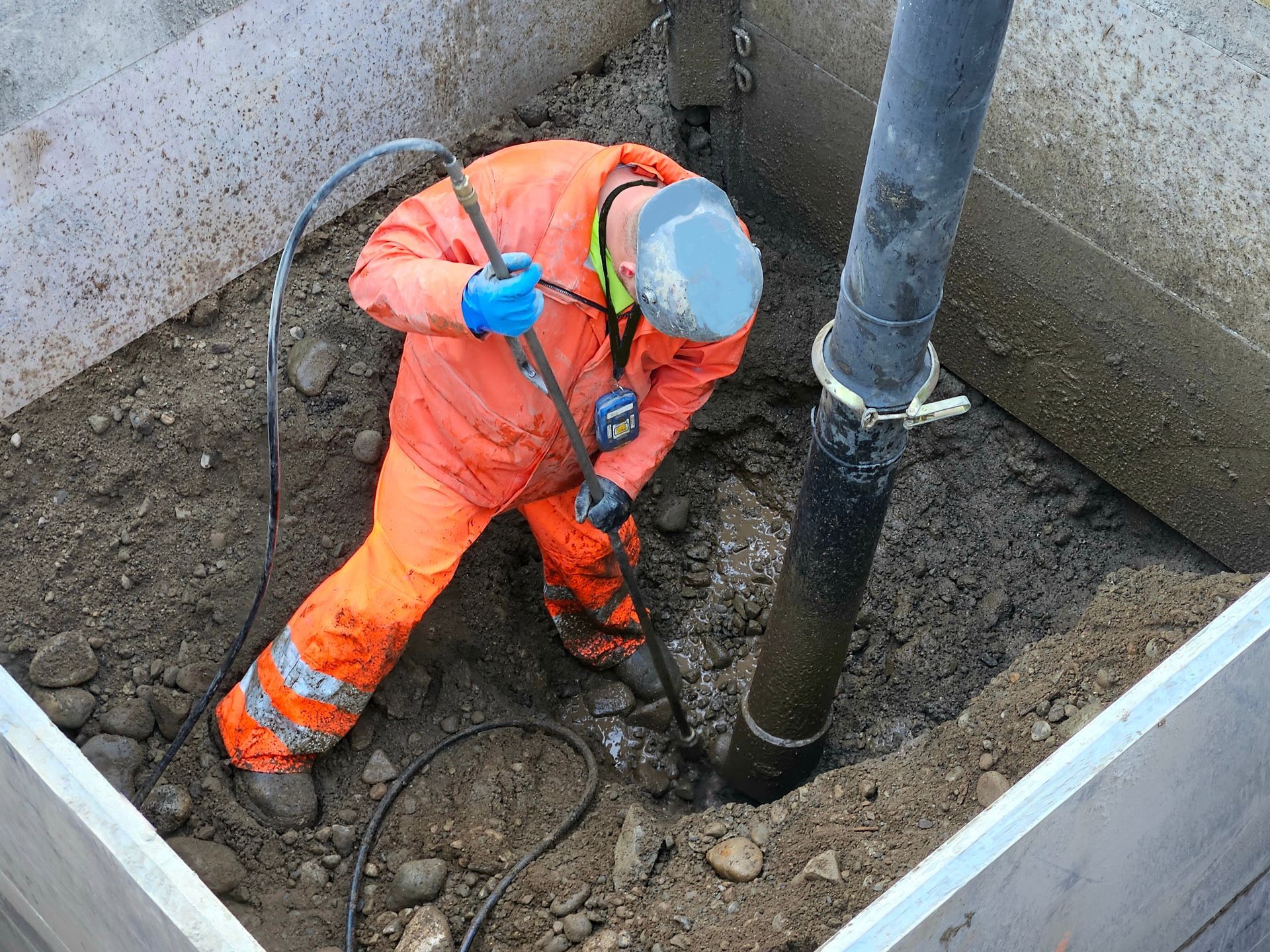 Worker in orange overalls using a tool near a large pipe in an excavation pit
