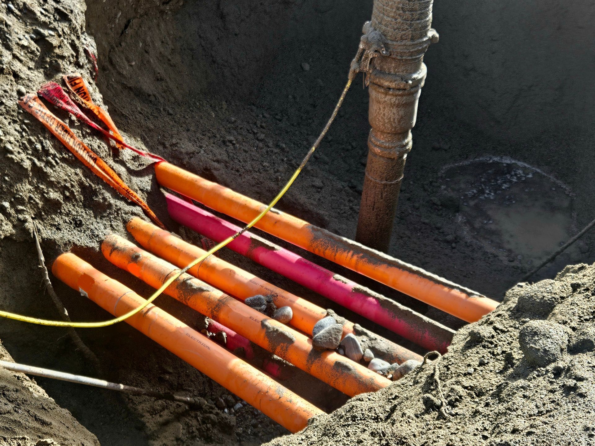 Orange and red utility conduits in an excavated hole next to a brown pipe