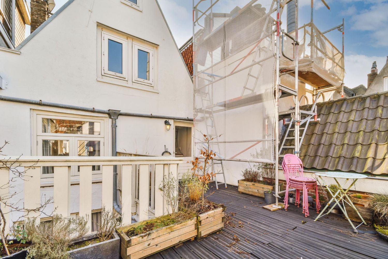 Balcony of a white building with wooden railings, pink chair, plants, and scaffolding on a roof.