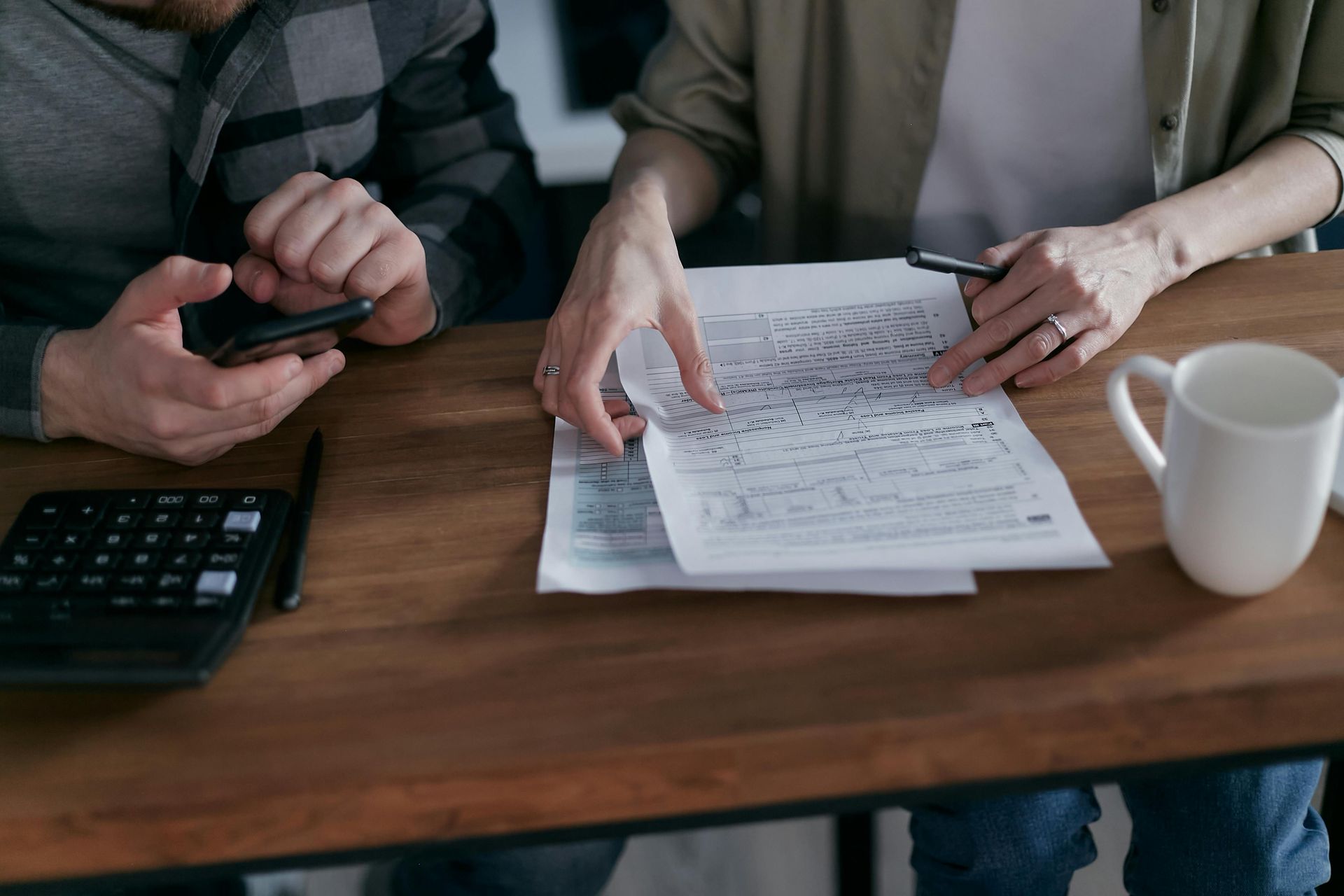 Couple reviewing paperwork with calculator, phone, and mug on a wooden table.