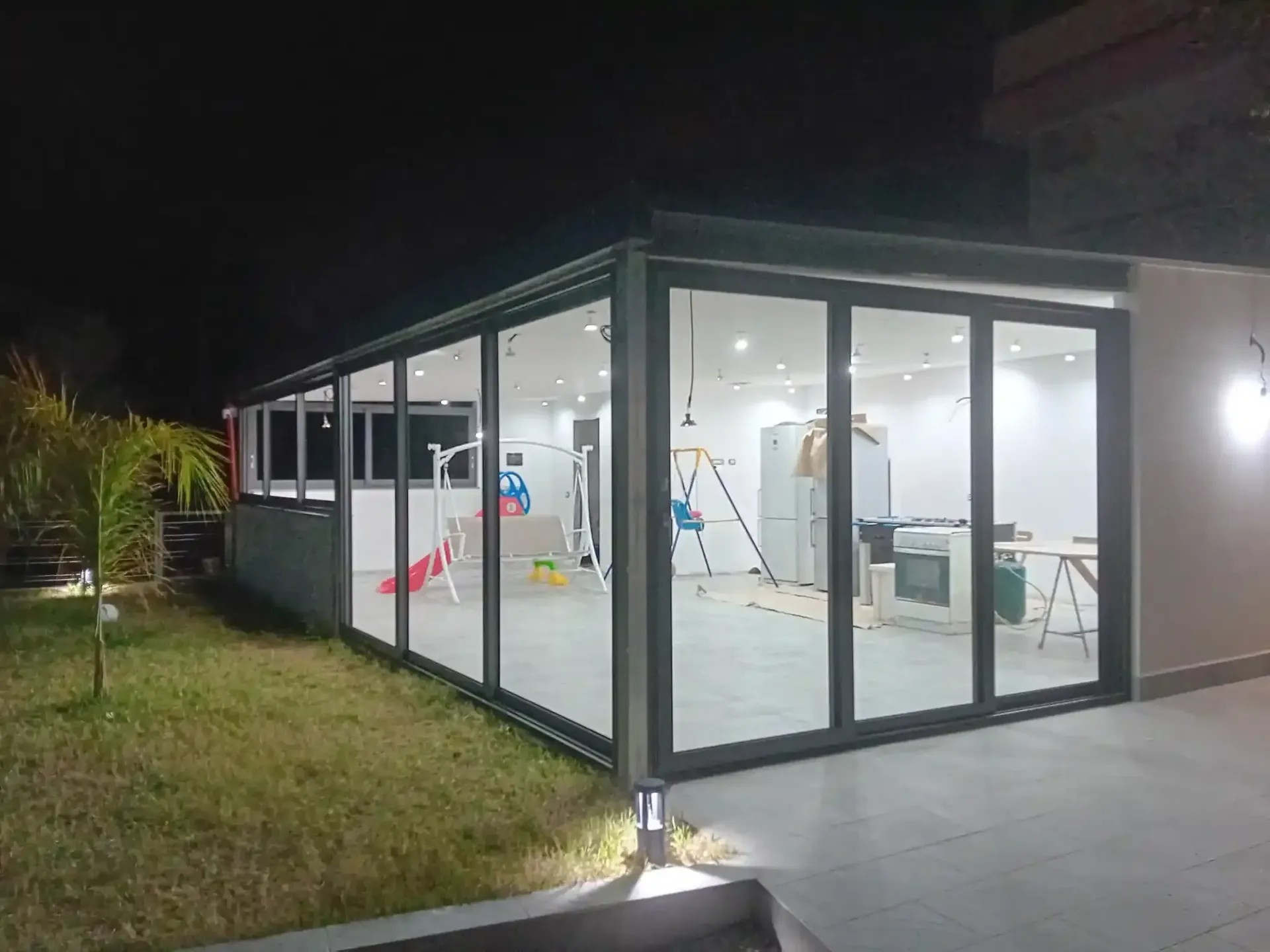 Veranda con pareti in vetro di notte, che illumina l'interno con luce bianca. L'esterno è caratterizzato da prato e patio.