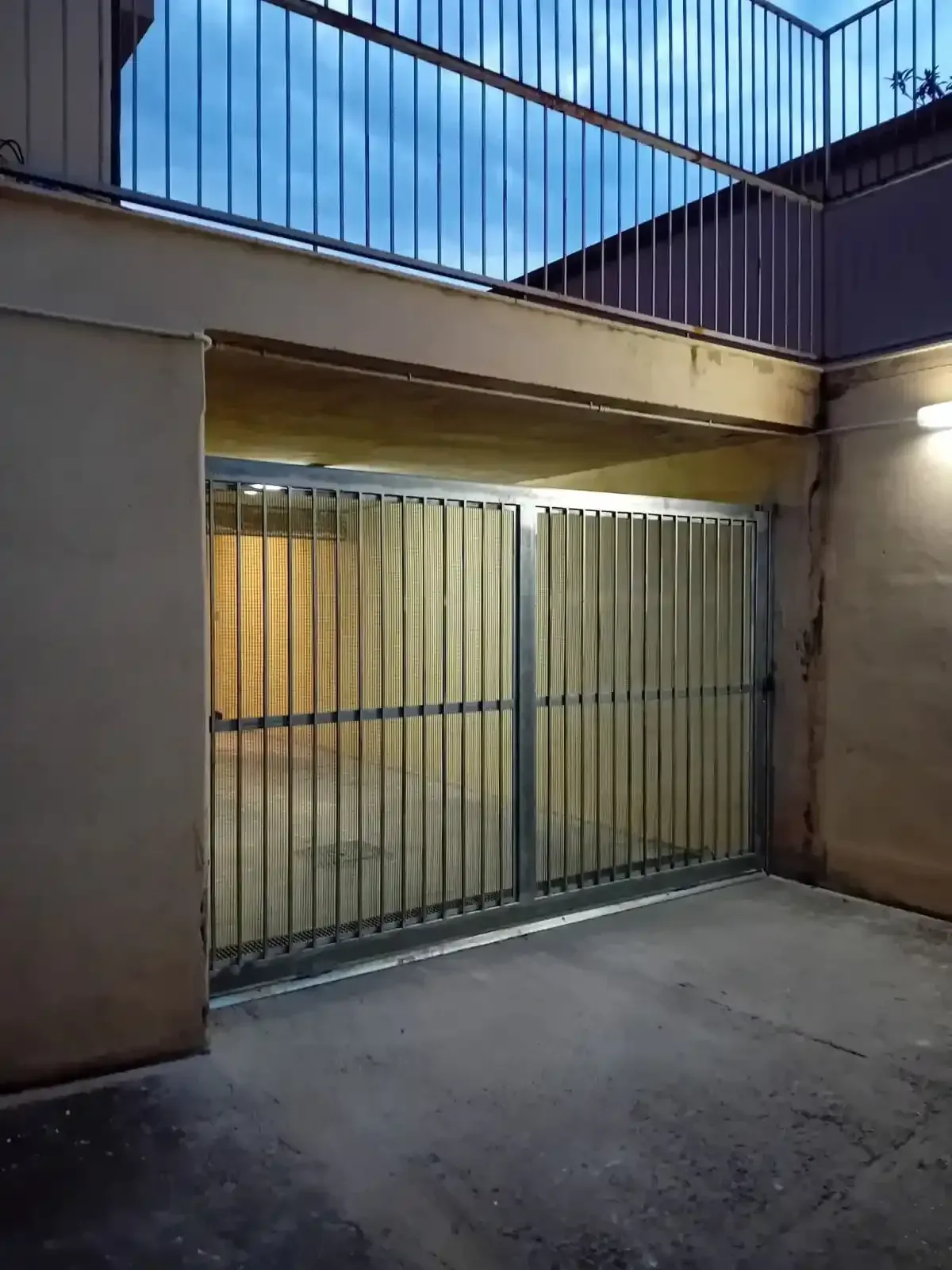 Cancelli metallici sotto una sporgenza di cemento con un balcone sopra, illuminati da una luce calda, esterno al crepuscolo.