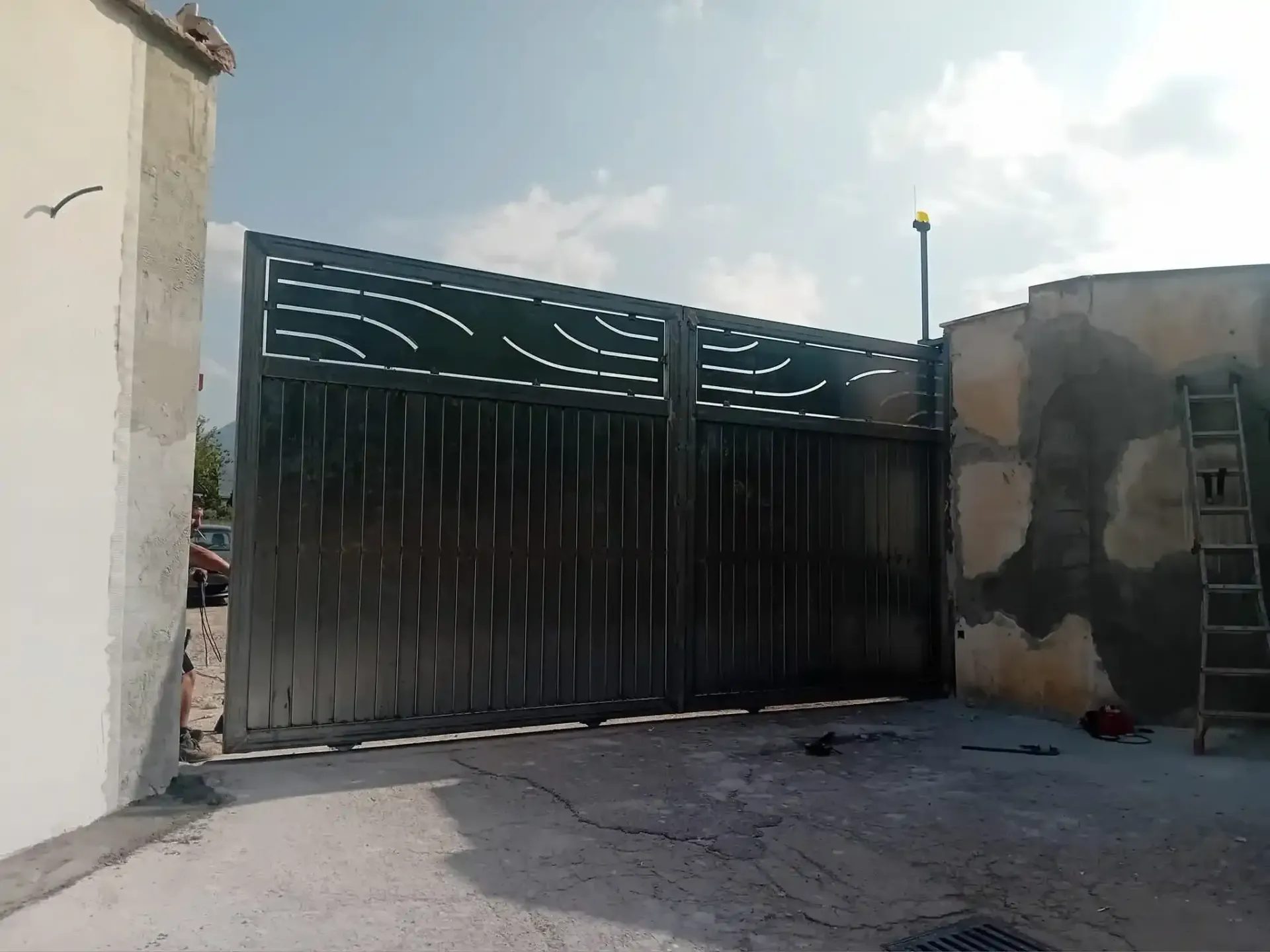 Cancello in metallo con parte superiore decorativa, tra muri di cemento, aperto su un'area pavimentata, scala a destra.