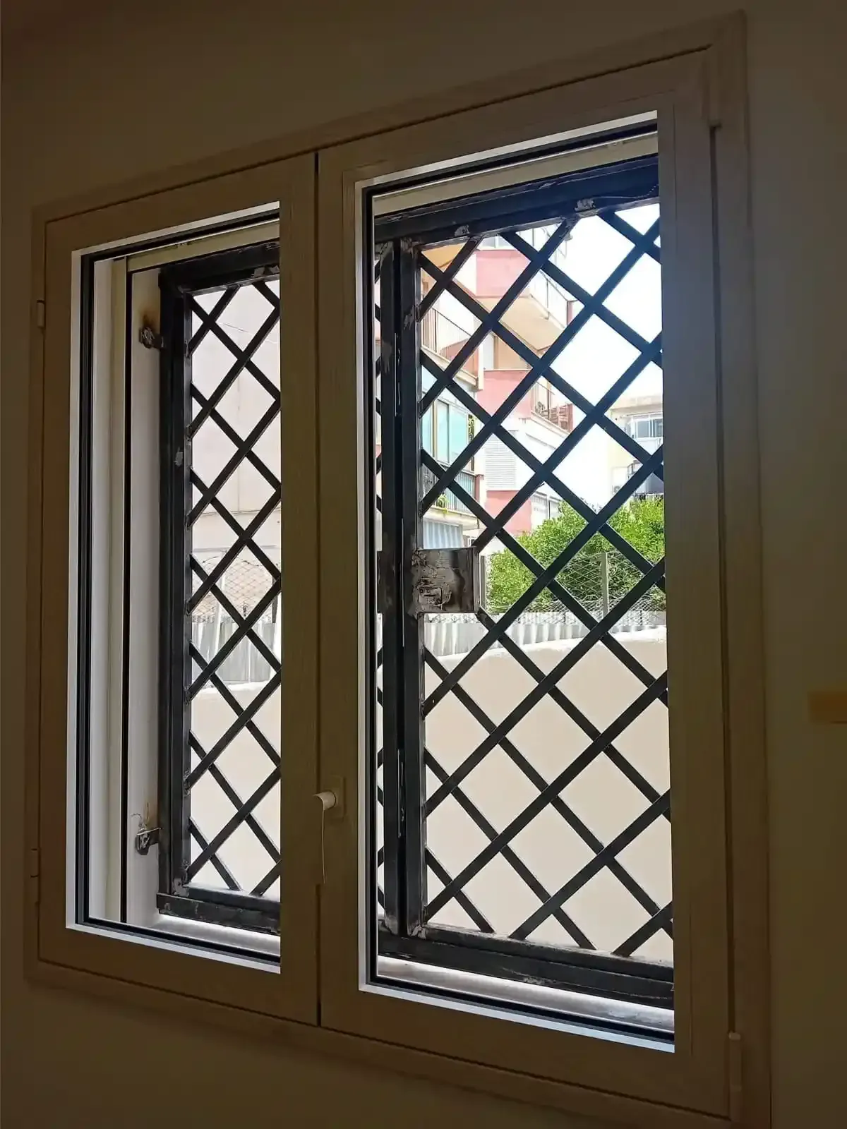 Due finestre con cornice bianca e grate di sicurezza nere a forma di diamante. La vista esterna mostra edifici e vegetazione.