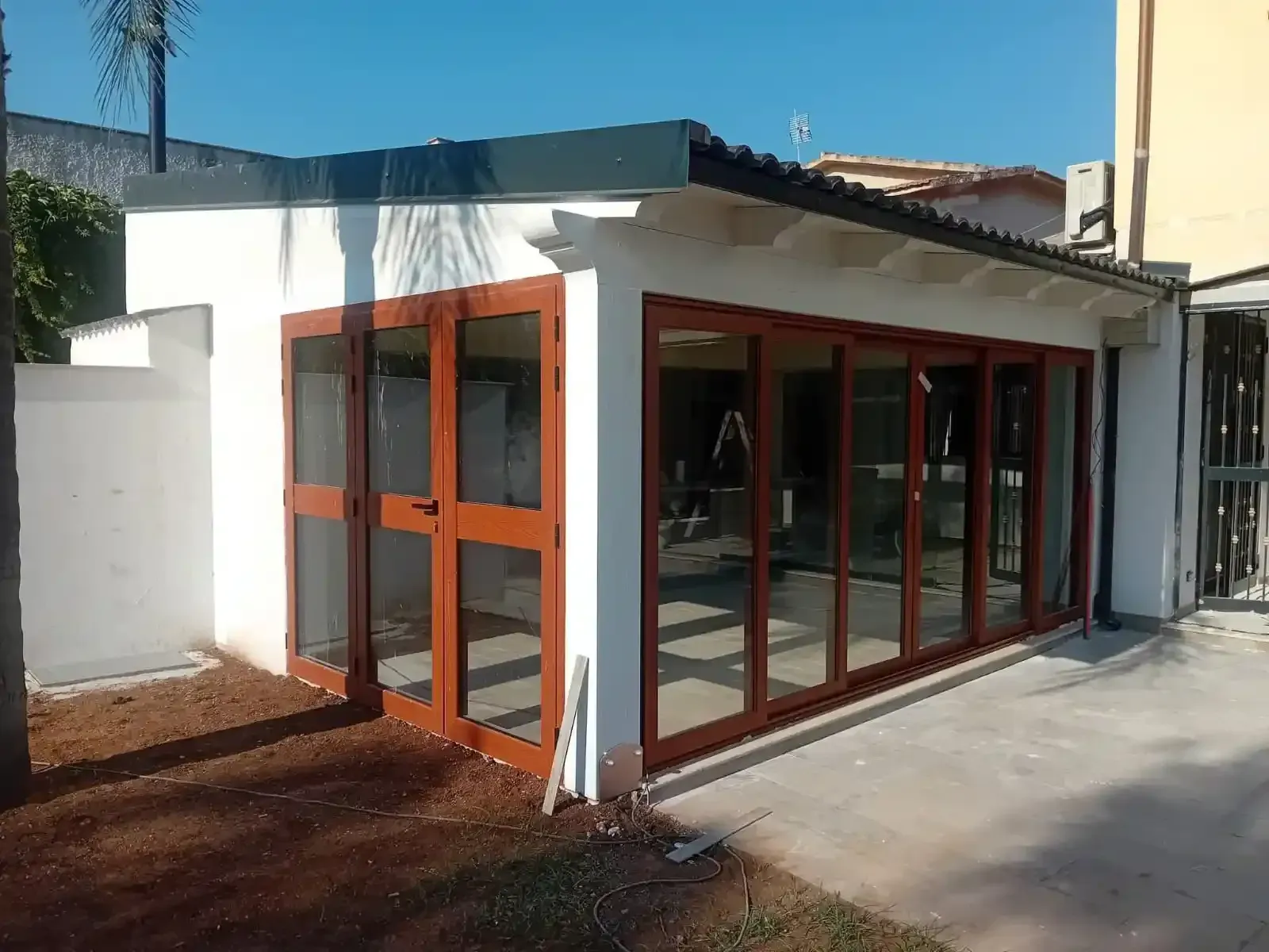Edificio bianco con porte e finestre in vetro con cornice in legno marrone, sotto un tetto scuro, in una giornata di sole.