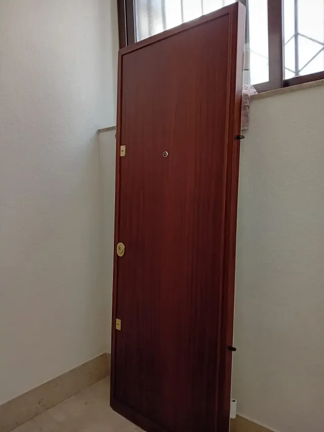 Una porta di legno marrone chiusa in un angolo contro le pareti bianche, con una piccola finestra visibile.