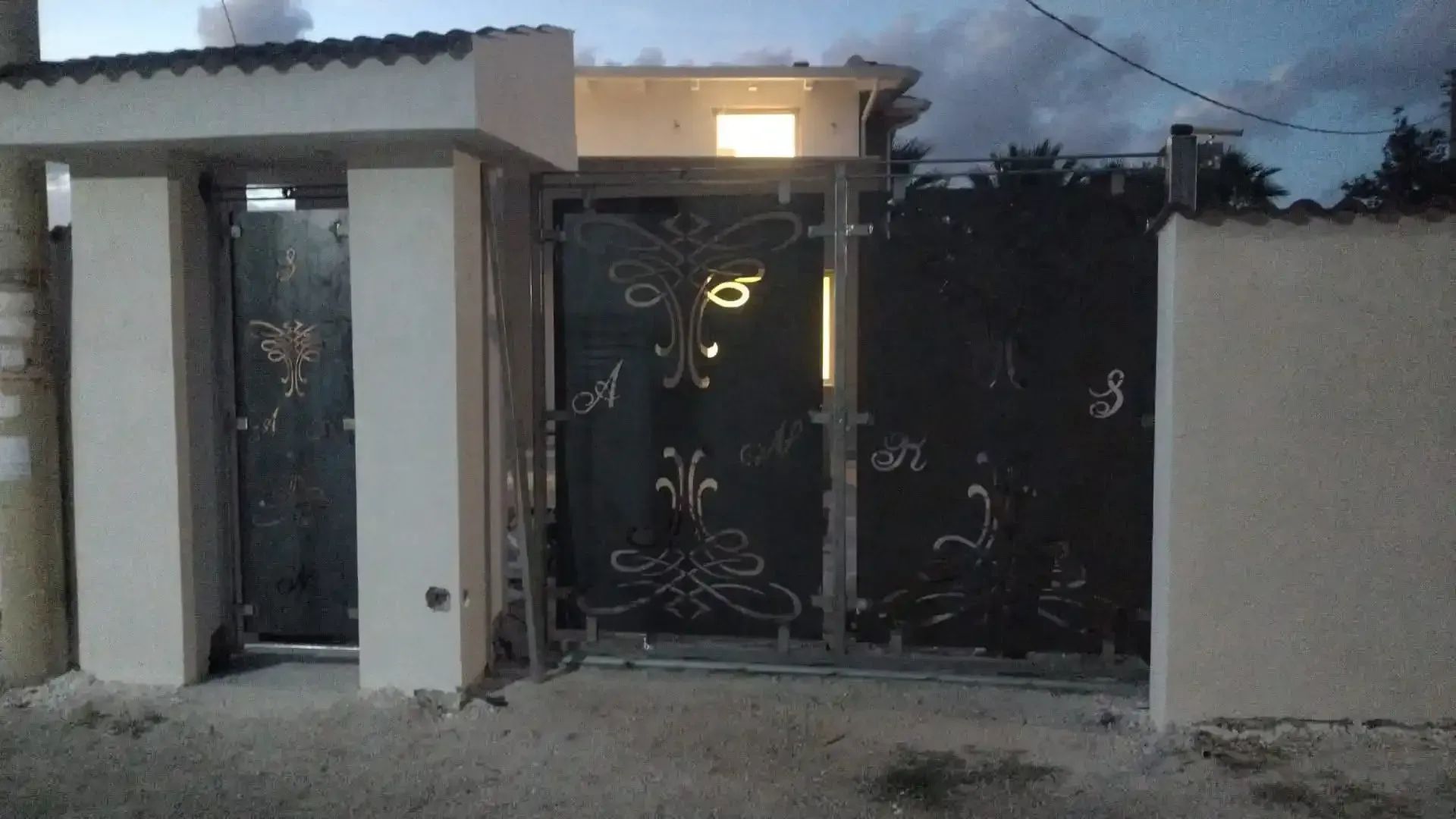 Cancello decorativo in metallo nero e pilastri bianchi su una strada al crepuscolo. Una finestra emette luce dall'alto.