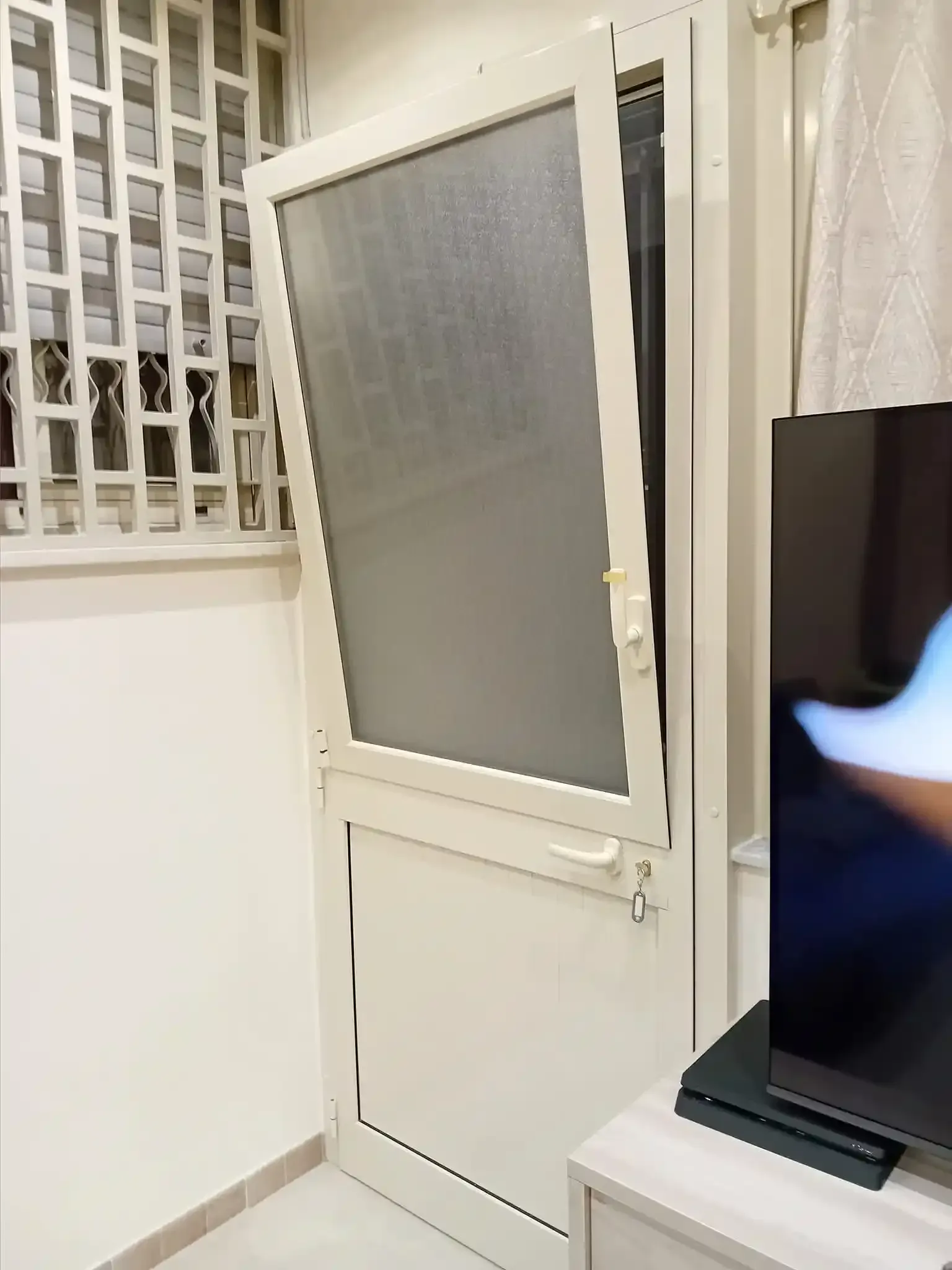 Porta a zanzariera bianca socchiusa, ad angolo contro una parete bianca. Finestra con griglia e TV visibili sullo sfondo.