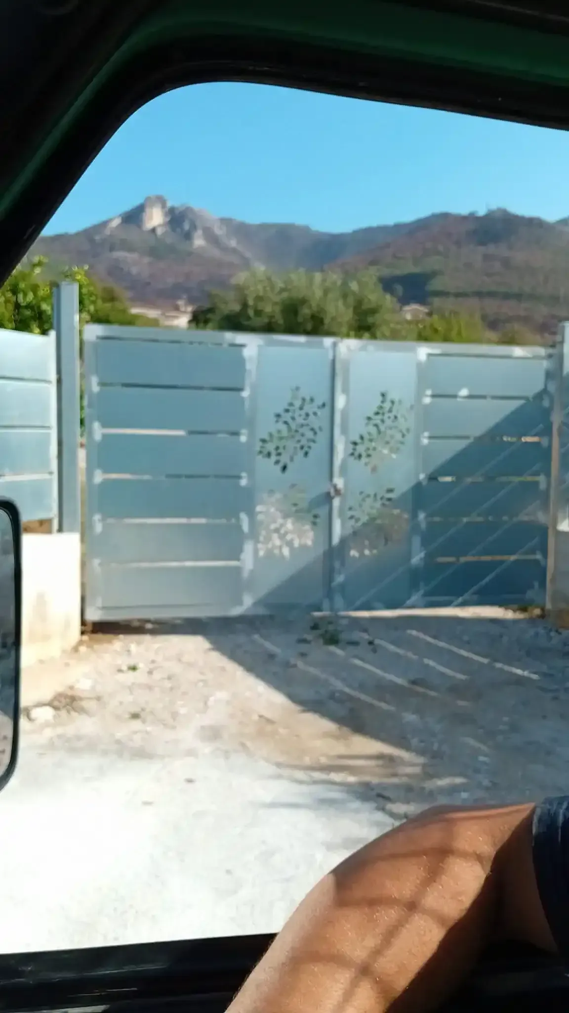 Cancello metallico con disegno di alberi, montagne sullo sfondo. La luce del sole proietta ombre.