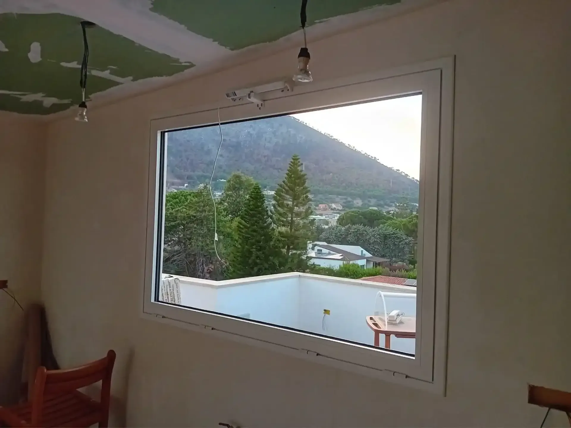 Finestra con vista su una montagna, alberi ed edifici; interno di una stanza con soffitto e pareti non finiti.