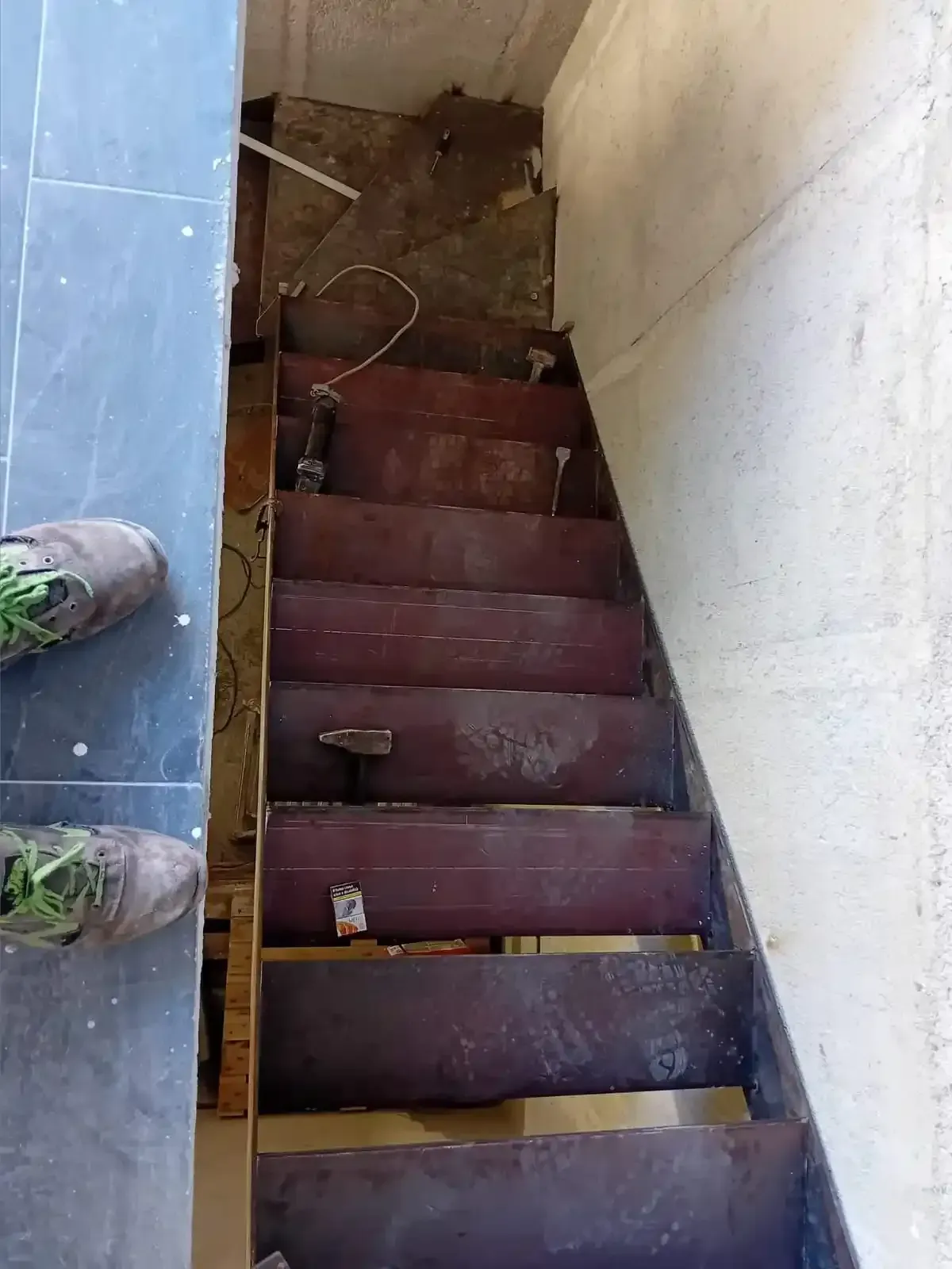 Scala in legno con gradini marrone scuro. I gradini sembrano grezzi. Sulla sinistra si vedono i piedi di una persona con gli stivali da lavoro.