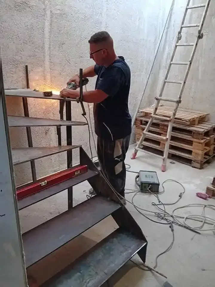 Un uomo salda una scala di metallo in una stanza; nelle vicinanze ci sono una scala e dei pallet.