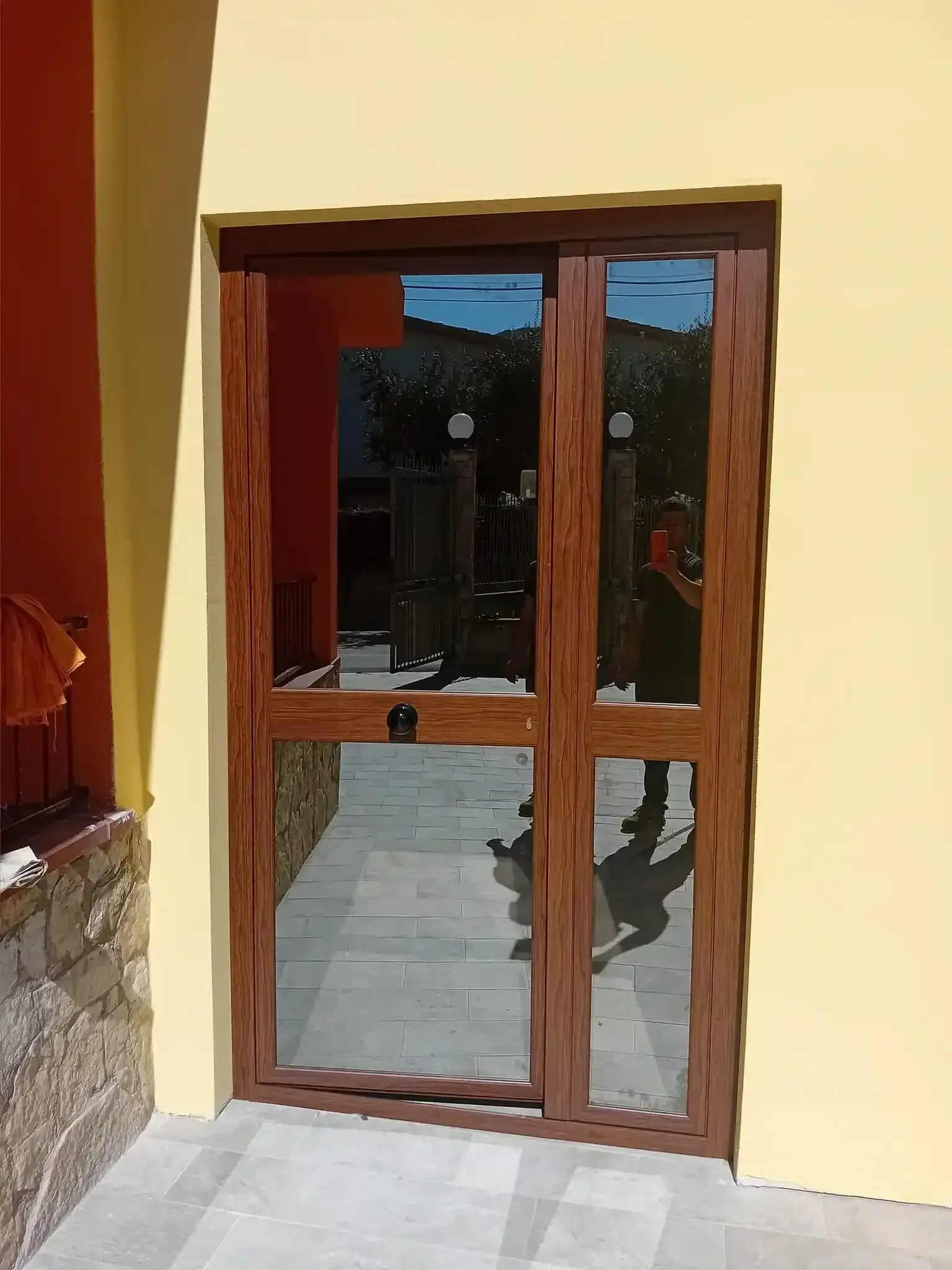 Porta in vetro con cornice marrone su una parete gialla, su cui si riflette una persona e un passaggio esterno.