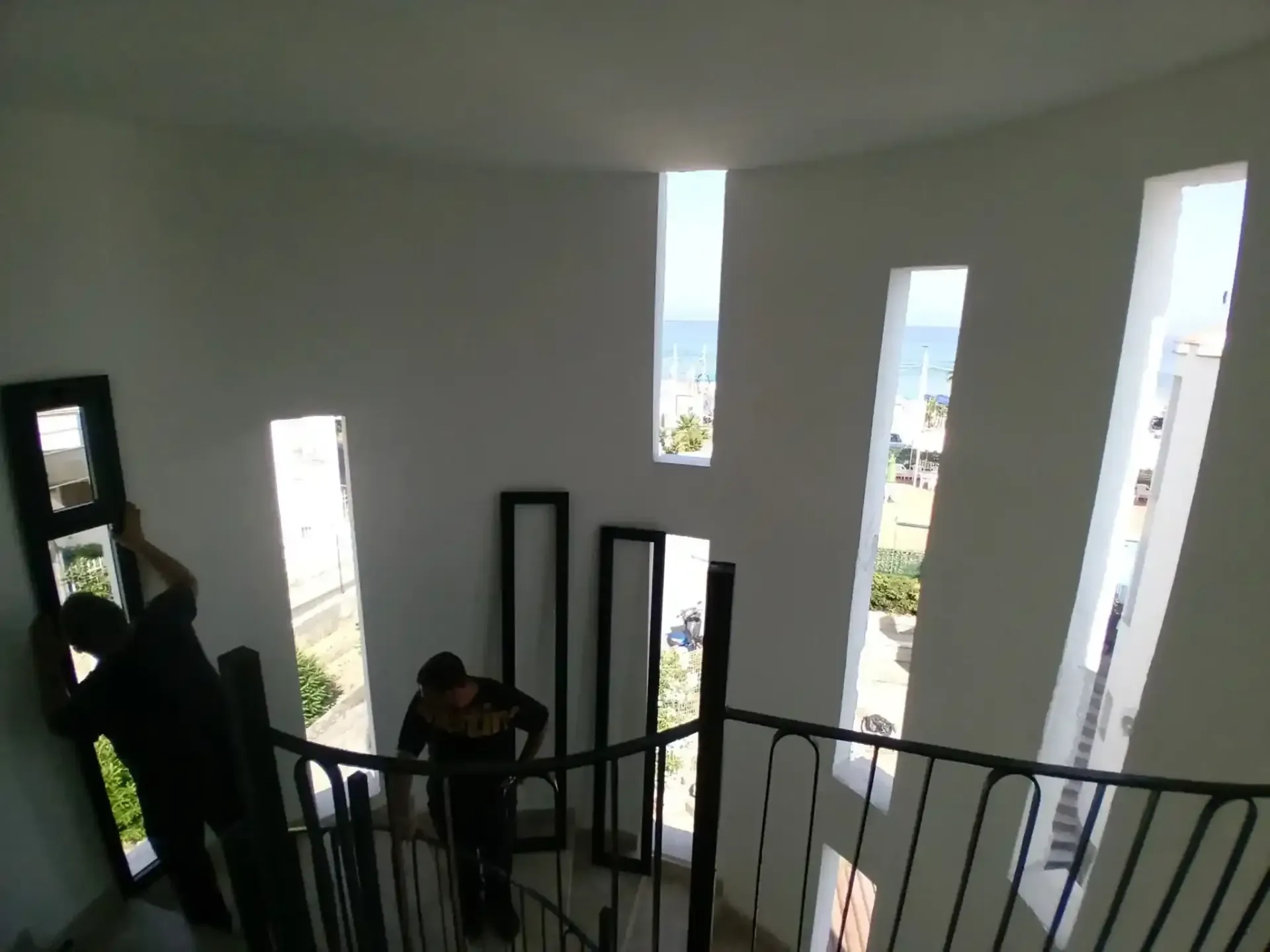 Due persone installano degli specchi alti all'interno di una scala a chiocciola bianca con vista sull'oceano.