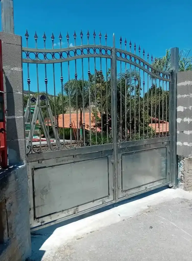 Cancello metallico con sommità arcuata e pinnacoli a punta, che conduce a un cortile con alberi ed edifici.