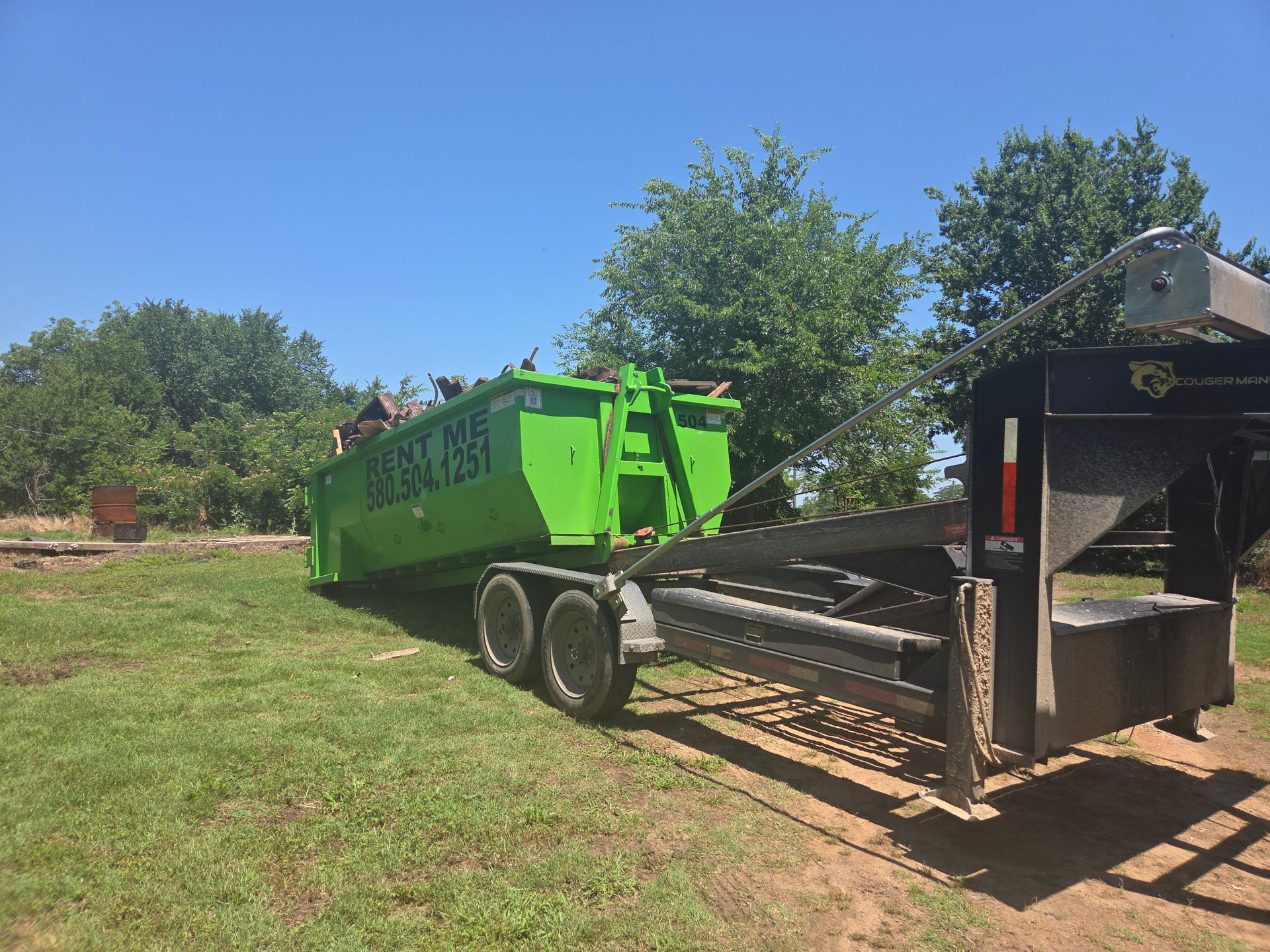Green dumpster on a trailer in a grassy area under a blue sky.