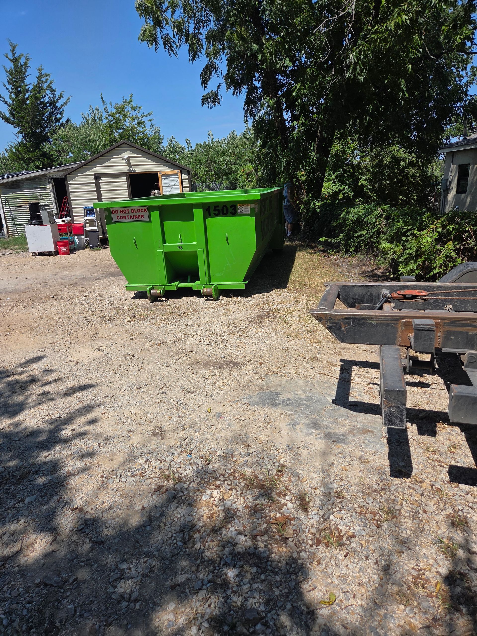 Bright green dumpster on gravel with small wooden buildings in the background.