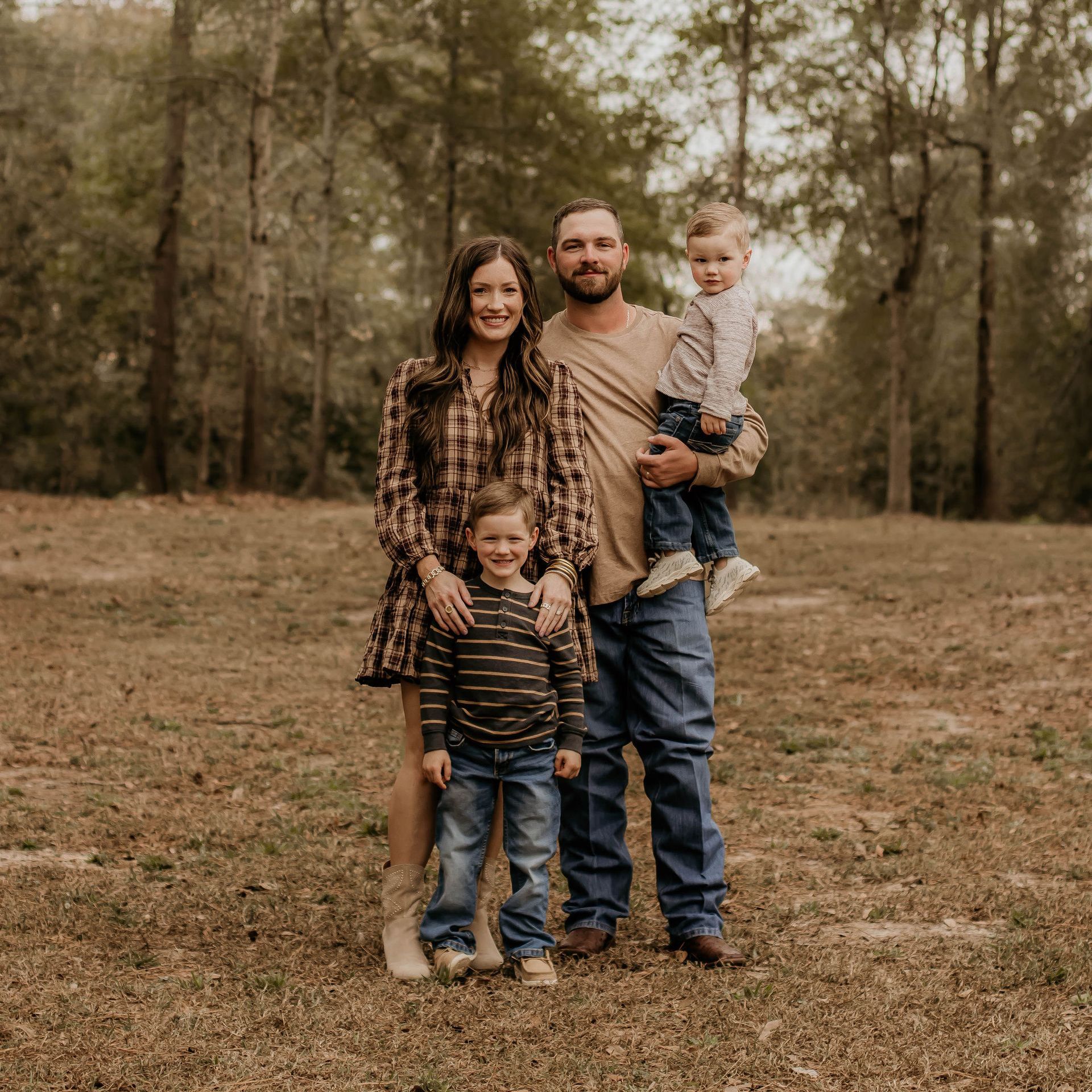 Family of four poses in a brown field with trees in the background.