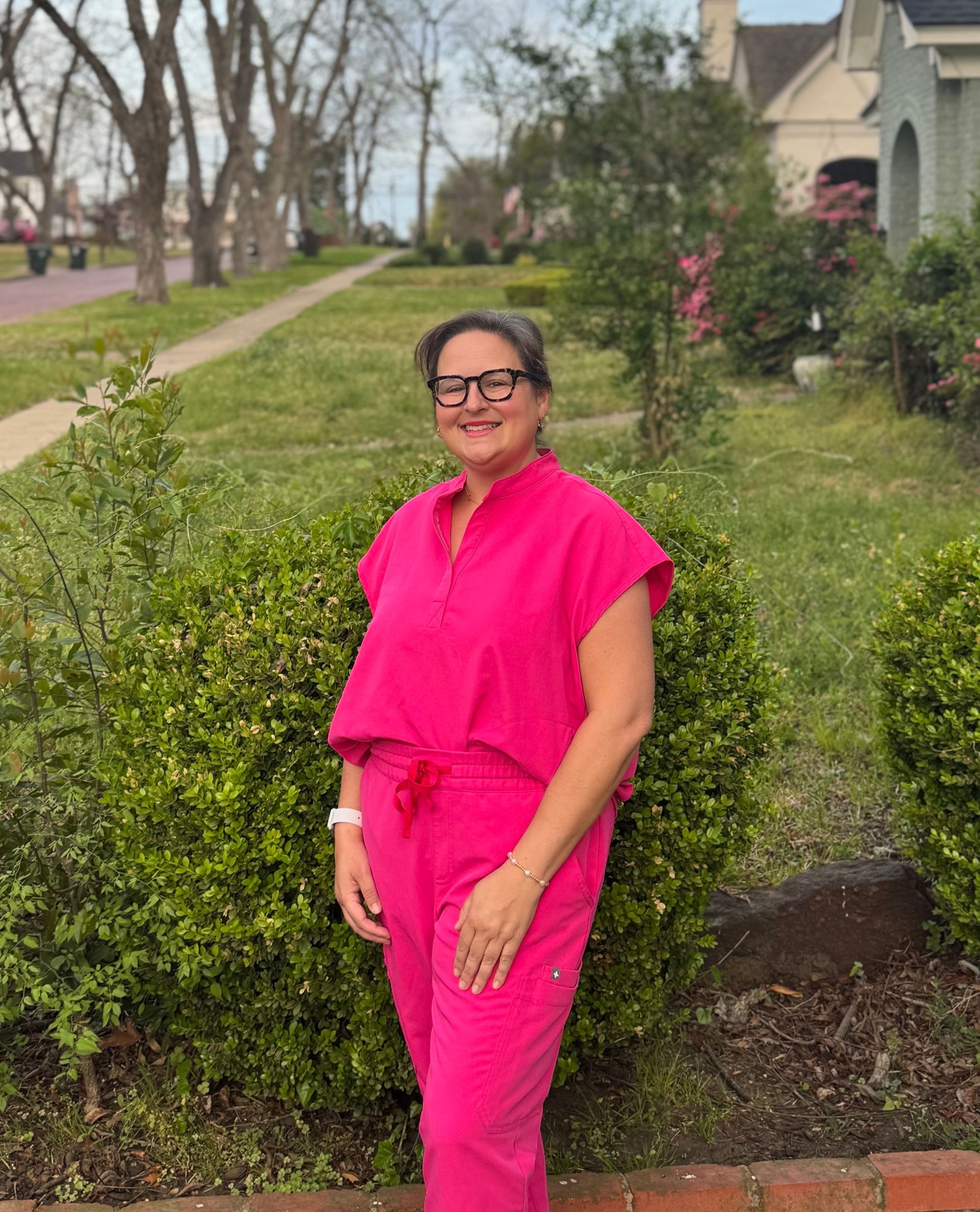 Woman in pink scrubs stands outside, smiles, near a green bush and a sidewalk in front of a house.