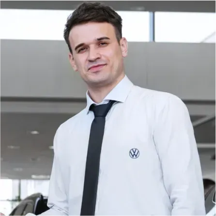Homem de camisa branca e gravata com o logotipo da Volkswagen, sorrindo em um showroom.