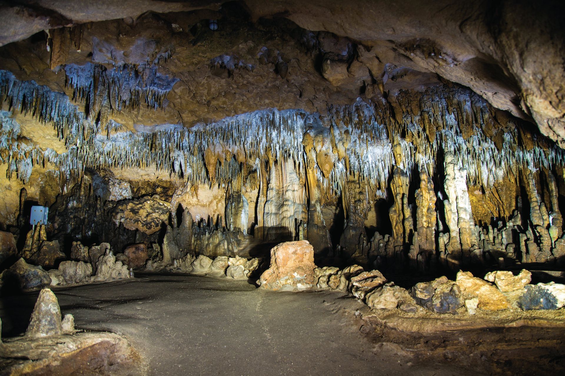 Inside of a large cave with stalactites and stalagmites lit with varying tones of light.
