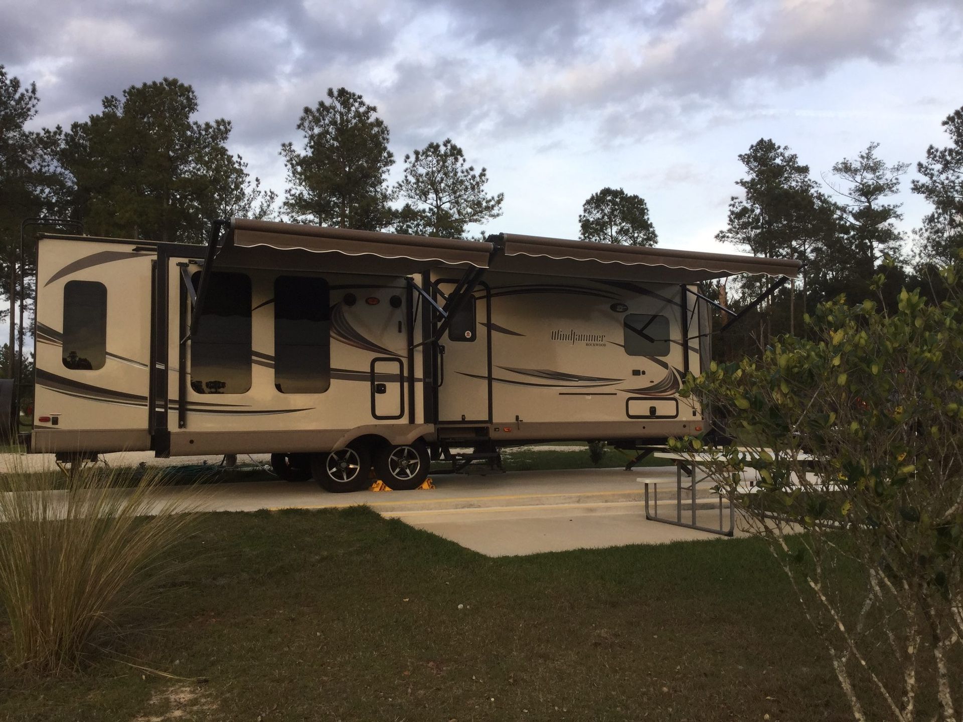 Tan RV parked in a campsite with the awning extended, trees in the background.
