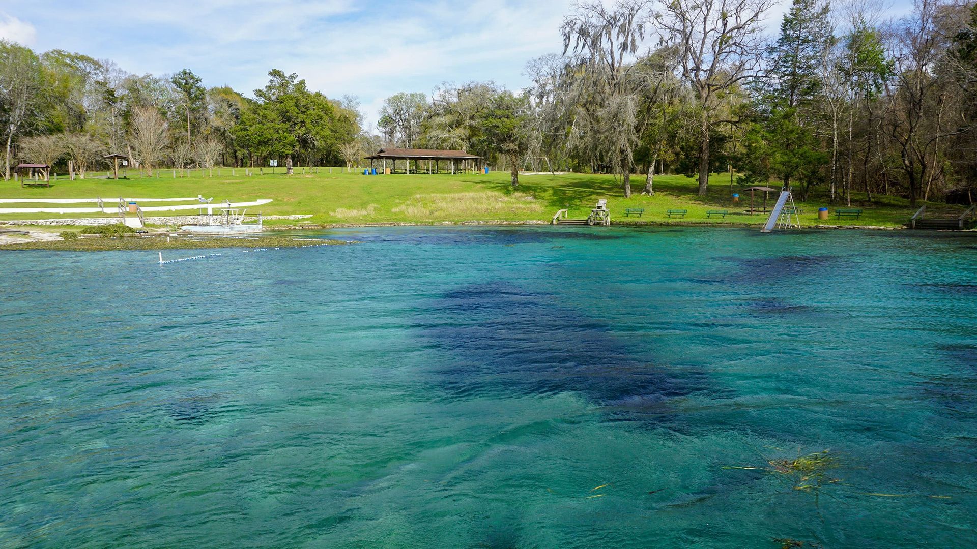 Clear turquoise spring water with grassy bank and trees; people near a gazebo.