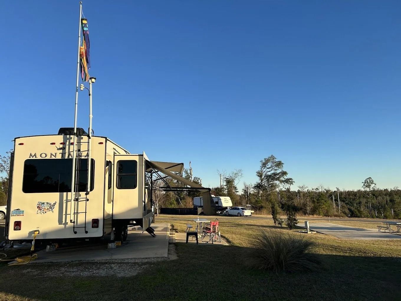 RV parked at a campsite, awning open. Blue sky overhead. Flags and a picnic table are visible.