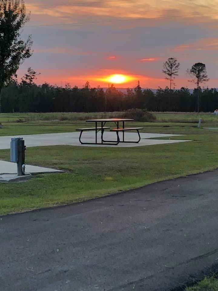 Large gray RV parked on a concrete pad at a campsite; a table and car are nearby.