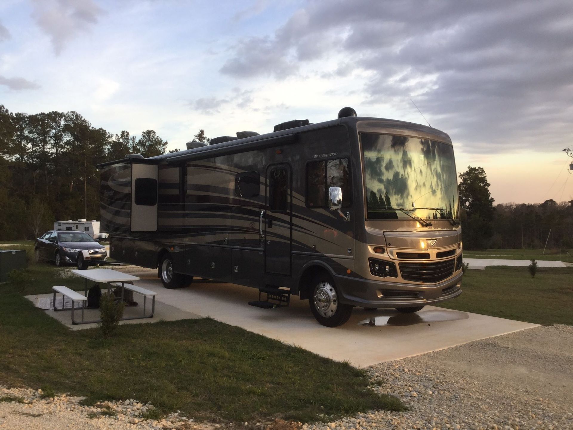 Large gray RV parked on a concrete pad at a campsite; a table and car are nearby.