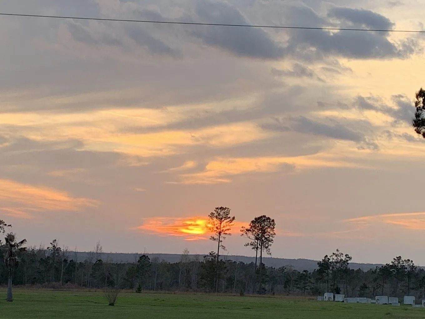 Sunset over a field and treeline, with orange and yellow hues in the clouds.