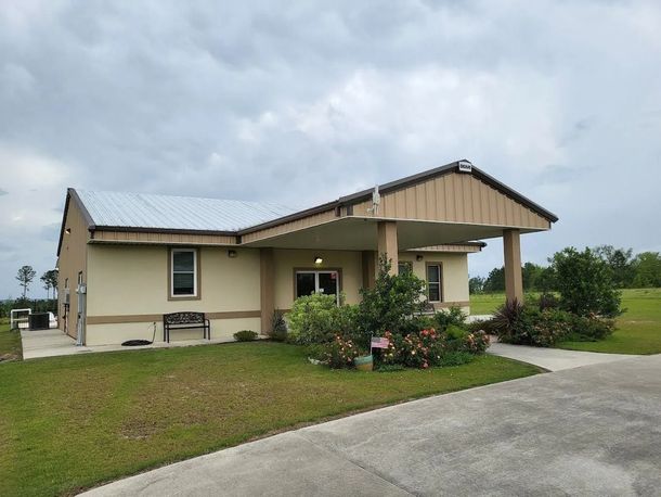 Beige building with a metal roof and a porch, surrounded by green grass. Cloudy sky.