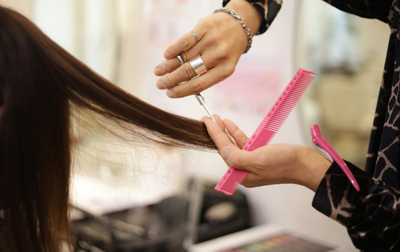 A hairstylist cuts long brown hair with scissors and a pink comb in a salon.