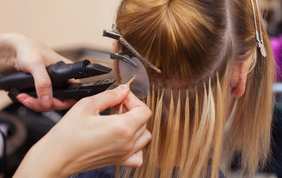 Hands attaching blonde hair extensions with a heat tool to the hair of a client.