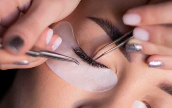 Eyelash extensions being applied; close-up of eye with under-eye patch, hands holding tweezers, white nails.