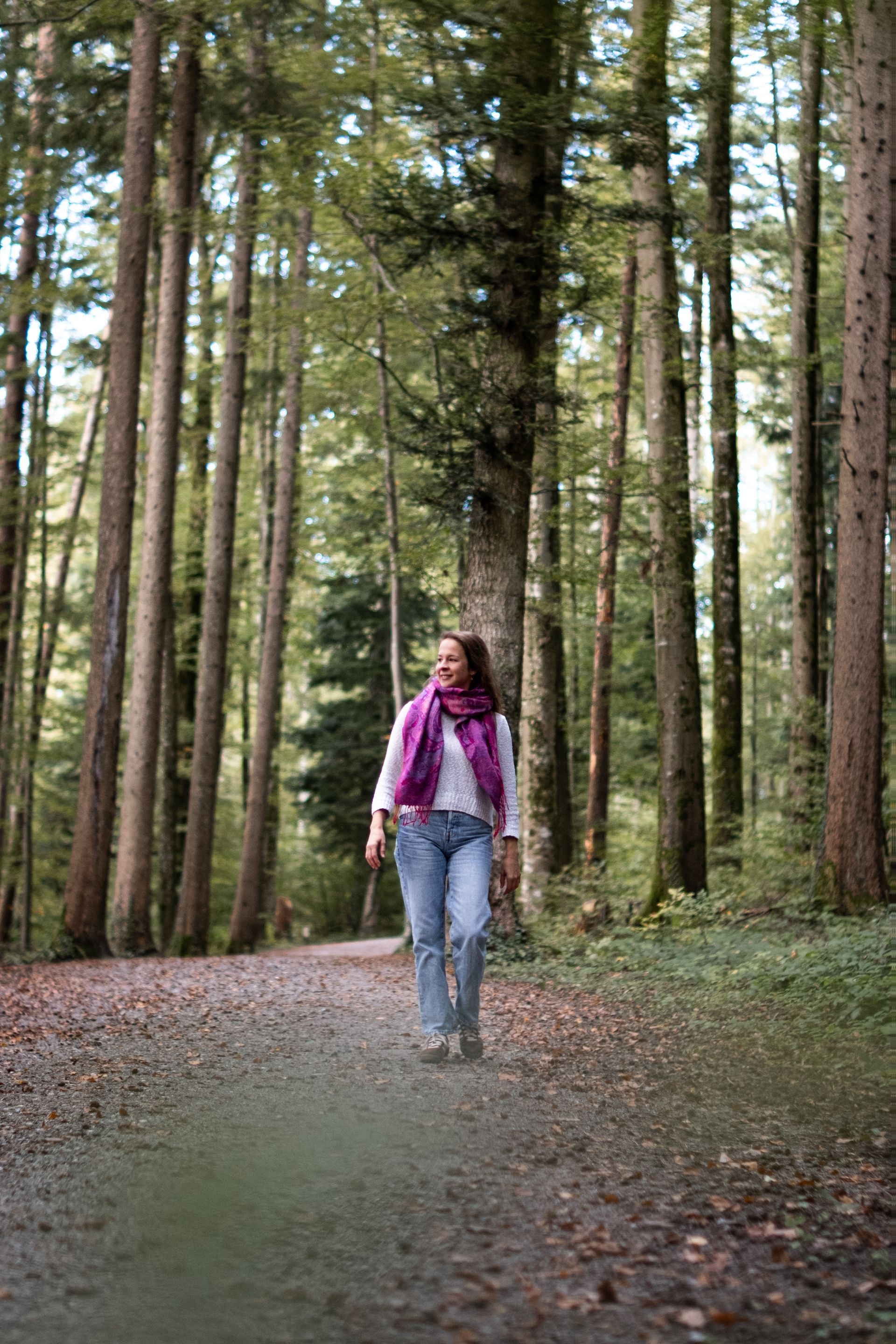 Eine Frau mit einem pinken Schal spaziert auf einem Weg durch einen Wald mit hohen Tannen.