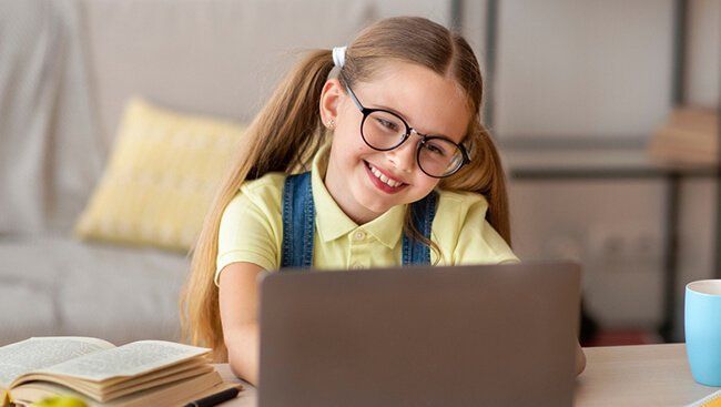 A Montessori child wearing glasses is sitting at a table using a laptop computer.