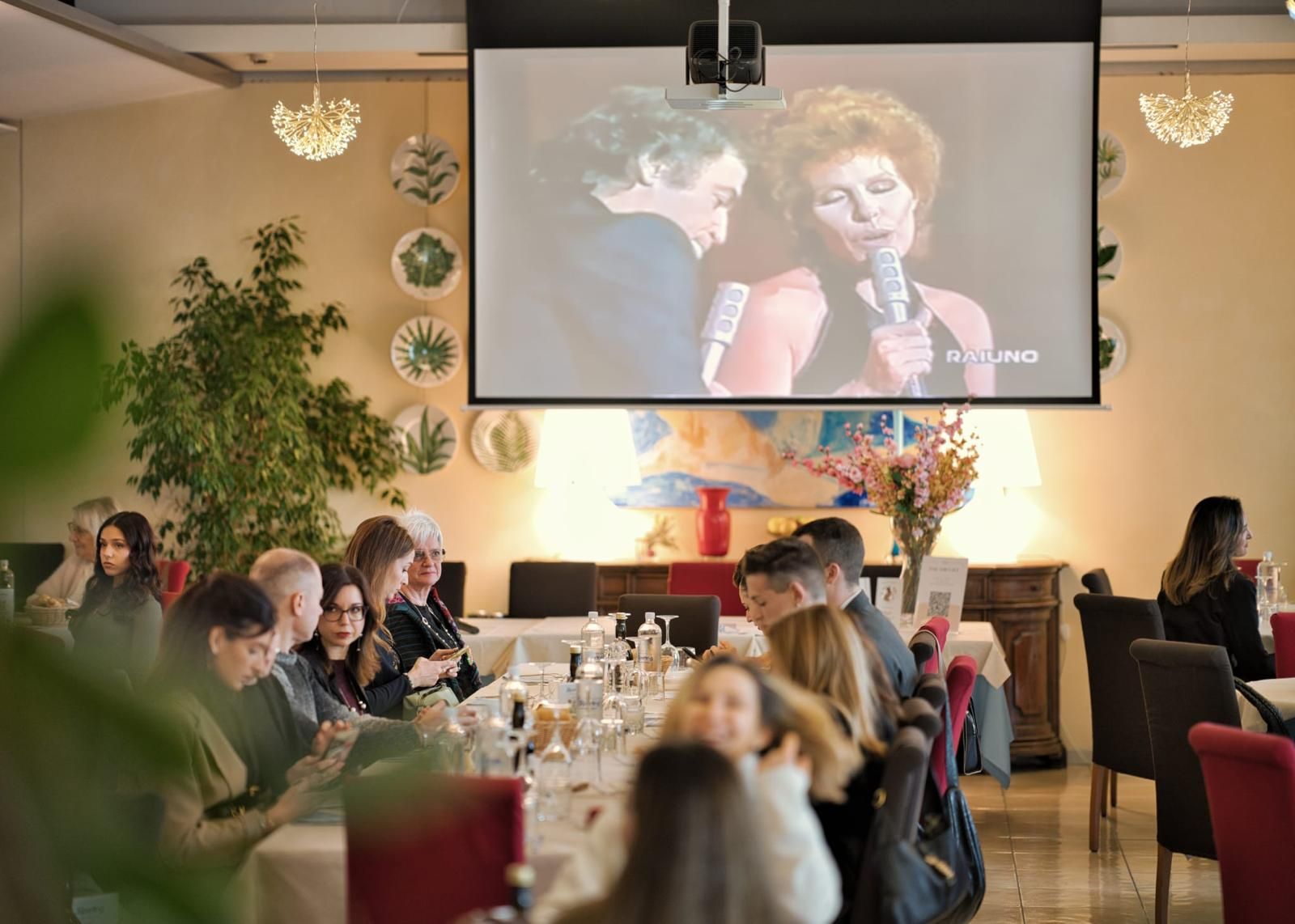 Interno di un ristorante, tavoli con persone che cenano. Tovaglie bianche, pareti dai toni neutri, luci decorative e finestre.