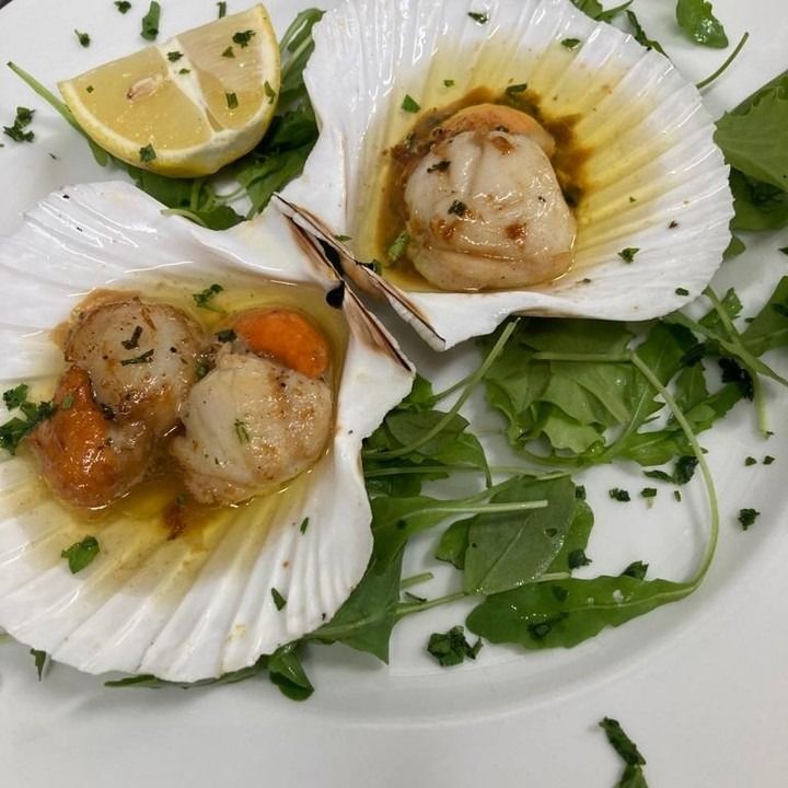 Capesante scottate nel guscio, guarnite con erbe aromatiche e spicchio di limone su un piatto.