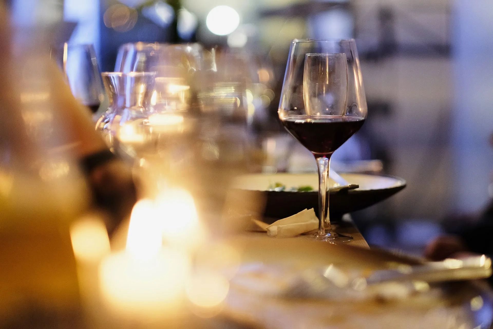 Bicchiere di vino rosso su un bancone scarsamente illuminato, con candela e piatti sullo sfondo.